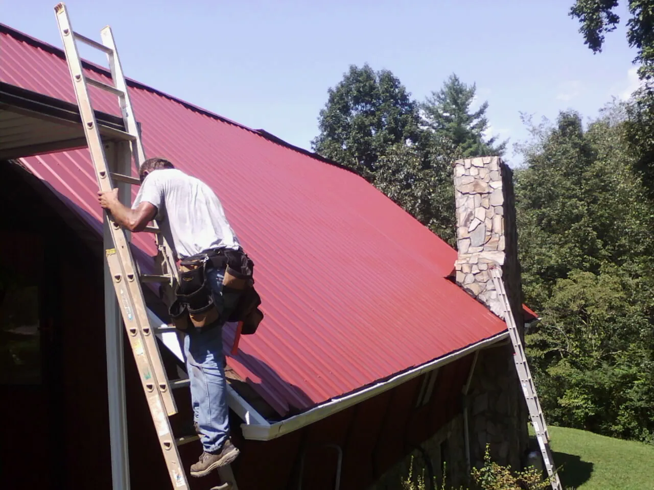 Roofer on a ladder beside a red metal roof and stone chimney — Soffit & Fascia Repair by Roofing Brothers in Colton